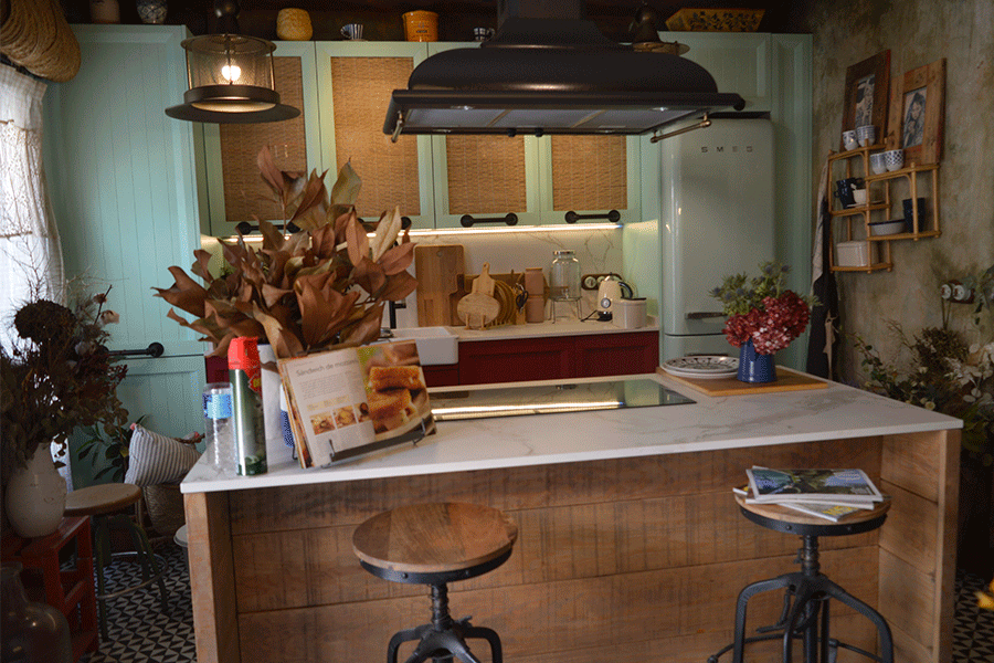 La cocina con isla central, papeles geométricos, muebles verdes y detalles rústicos.